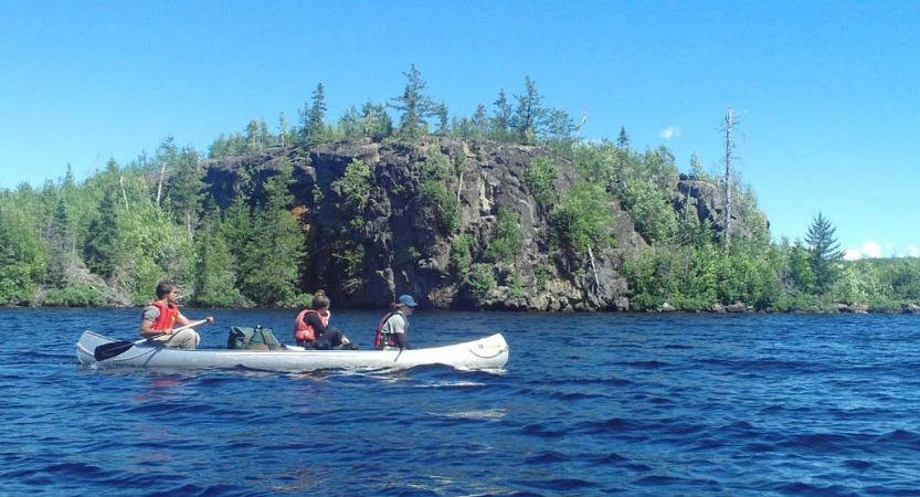 Three people paddle a canoe on blue water past a rocky shore.